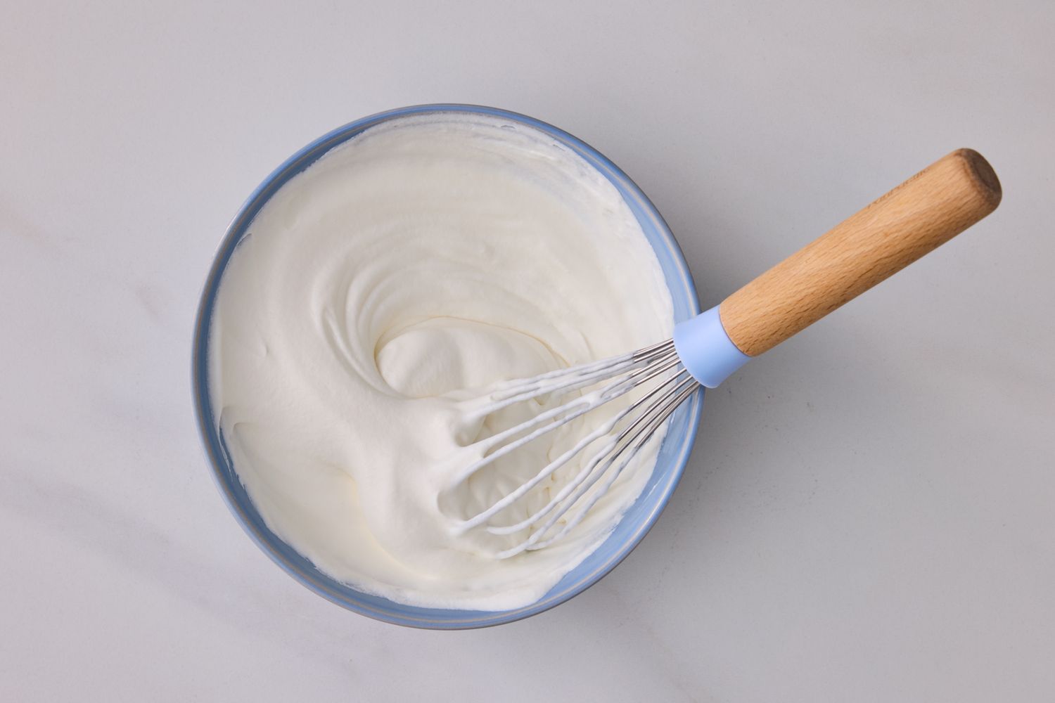 A bowl containing whipped cream placed on a surface with a whisk resting inside the bowl