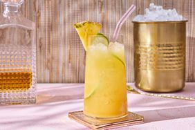 A pineapplerum cocktail in a glass with ice and garnished with lime and pineapple on a table with a decanter and an ice bucket