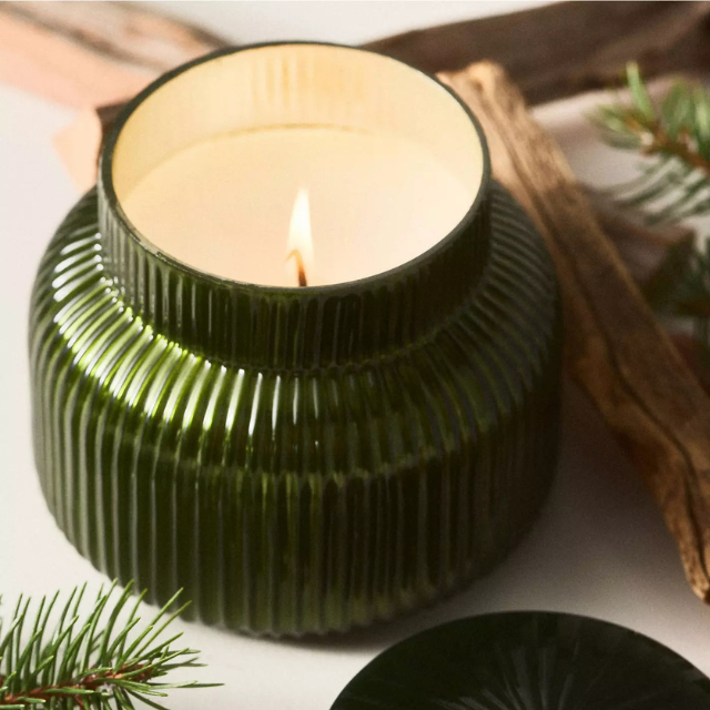 Lit candle in a textured green glass container with decorative branches and pine needles nearby