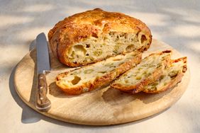 A loaf of bread with olives and cheese partially sliced on a wooden cutting board next to a knife