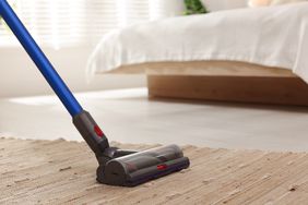 A vacuum cleaner being used on a woven rug a bed visible in the background