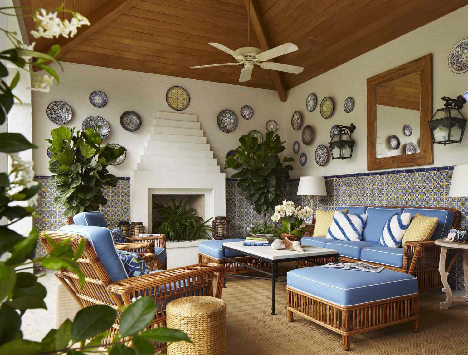 A cozy indoor patio with rattan furniture blue cushions decorative plates on the wall and green plants surrounding a white fireplace