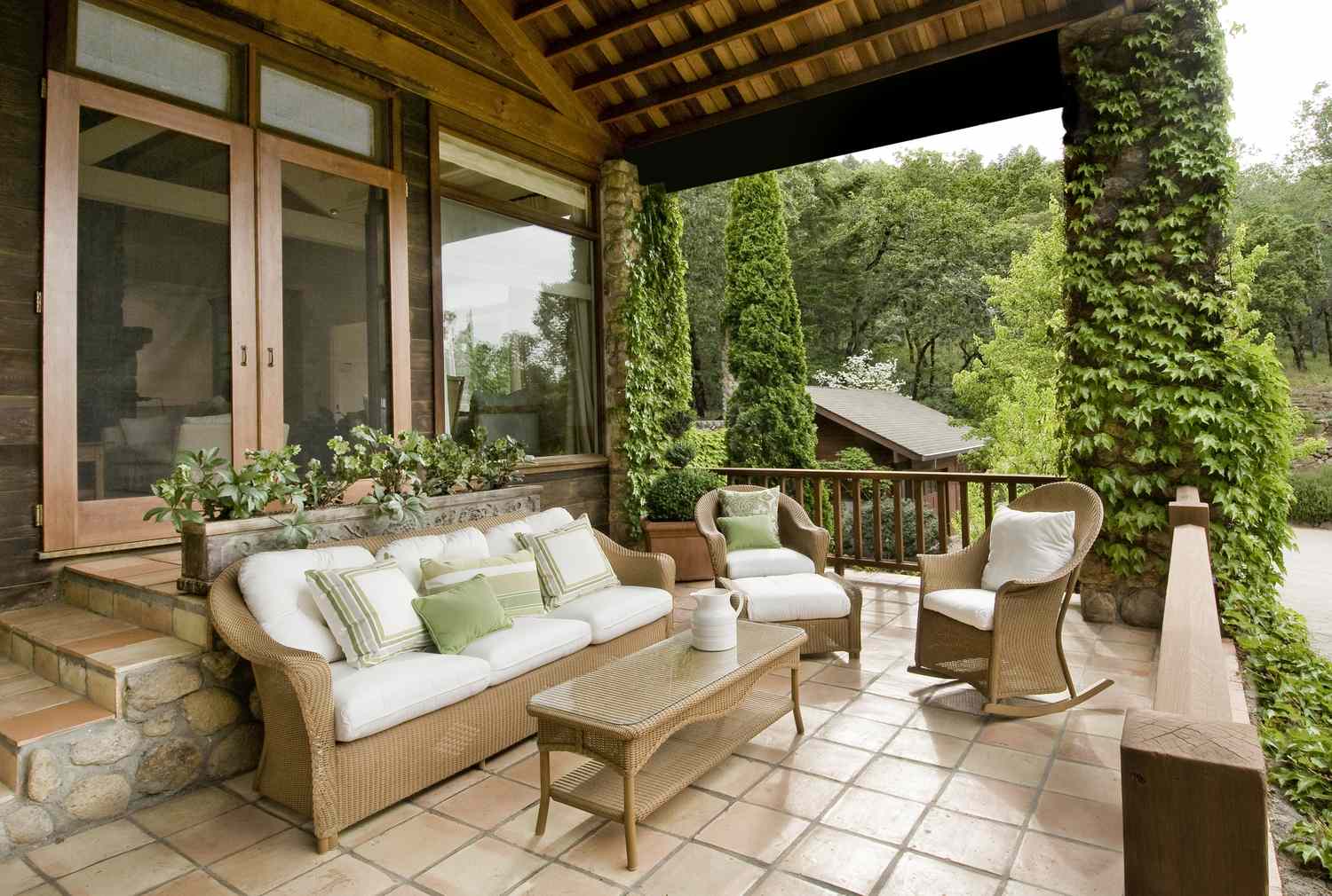 Outdoor patio with wicker furniture surrounded by greenery located next to a wooden house