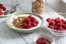 A bowl of yogurt garnished with granola and raspberries surrounded by a glass bowl of raspberries and jam on a wooden table