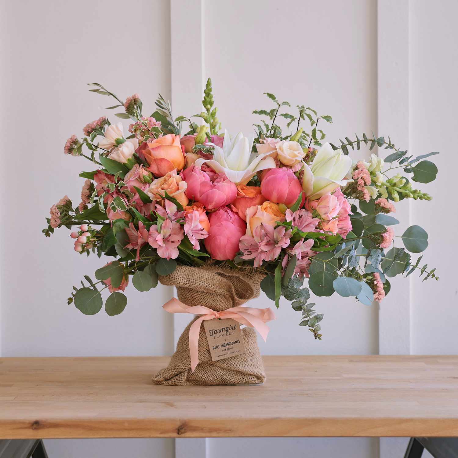 A bouquet of flowers wrapped in burlap and placed on a wooden table featuring peonies and other assorted blooms