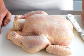 raw whole chicken on cutting board with knife and person in white
