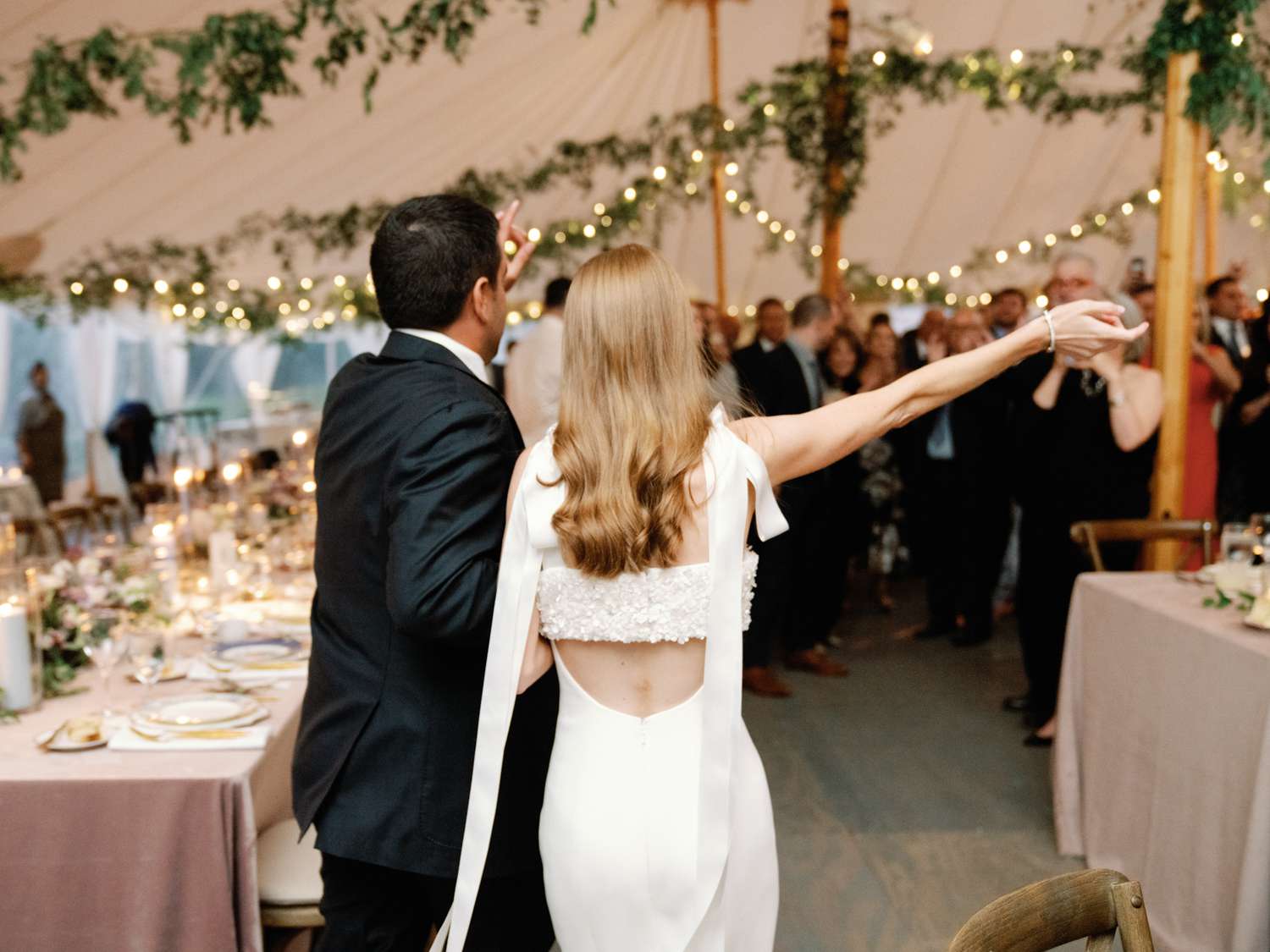 bride and groom walking into wedding reception tent embellished with draped lights and greenery
