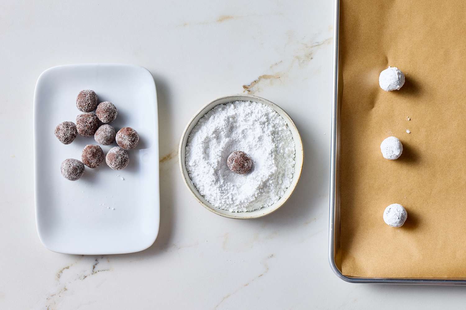 Cookie dough balls being coated in powdered sugar and placed on a baking tray lined with parchment paper