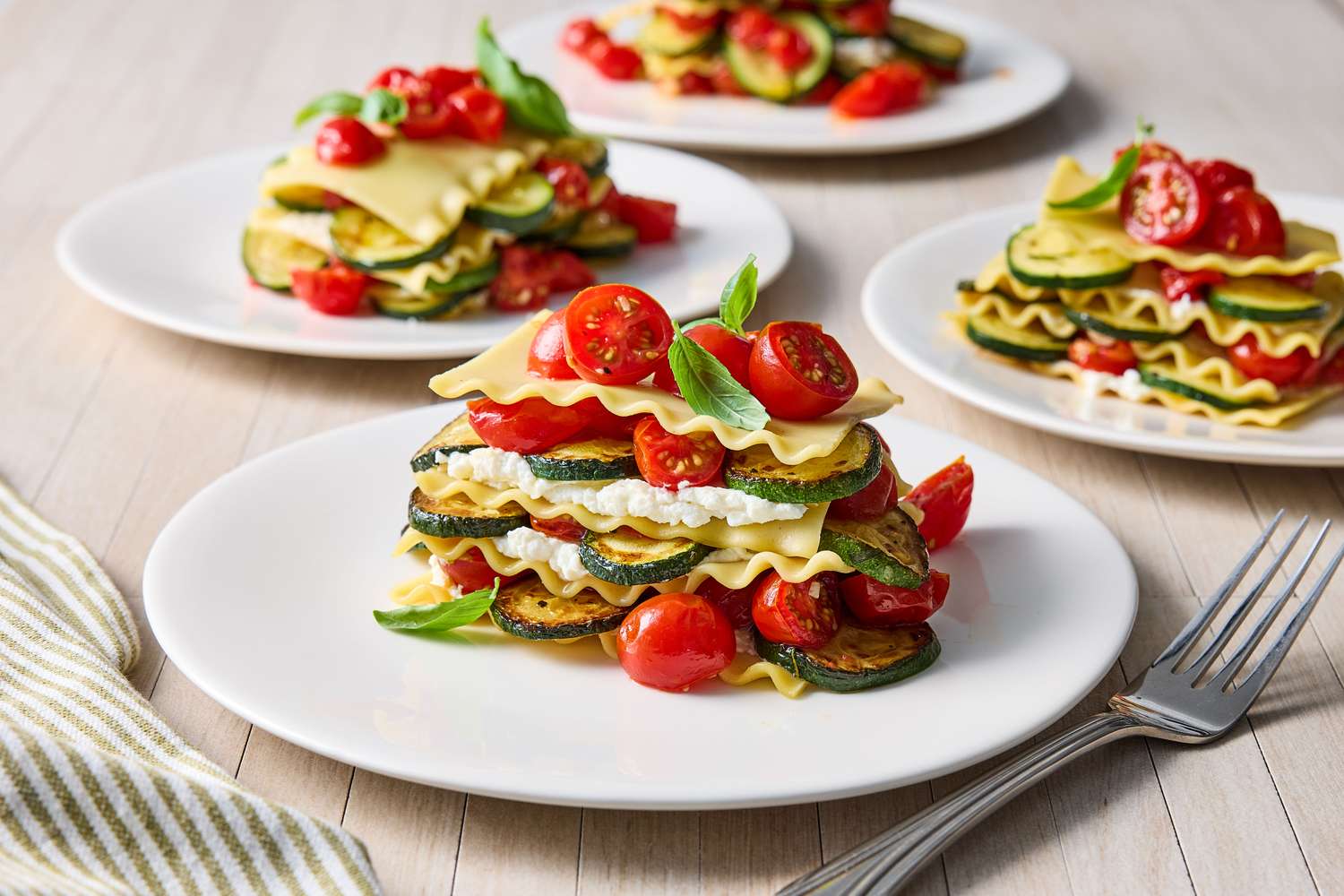 Layered dishes featuring slices of vegetables pasta sheets and ricotta cheese on white plates garnished with fresh ingredients