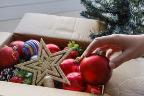 A hand reaching for a red bauble among various holiday ornaments in a box with a partially visible Christmas tree nearby