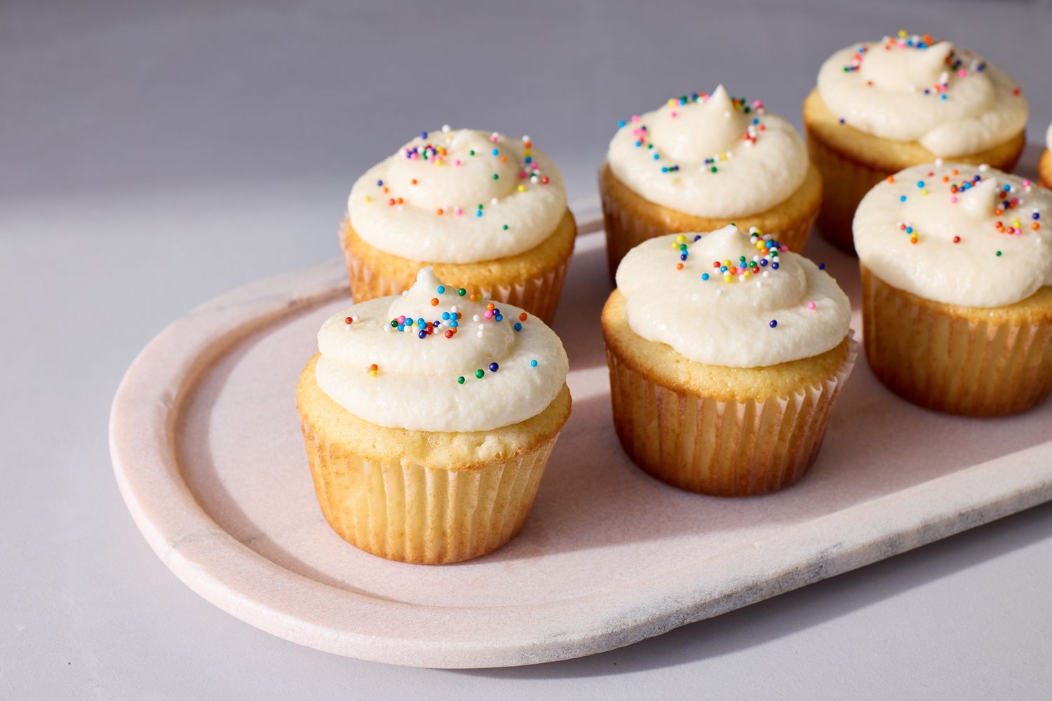 Six vanilla cupcakes with frosting and sprinkles arranged on a white oval plate