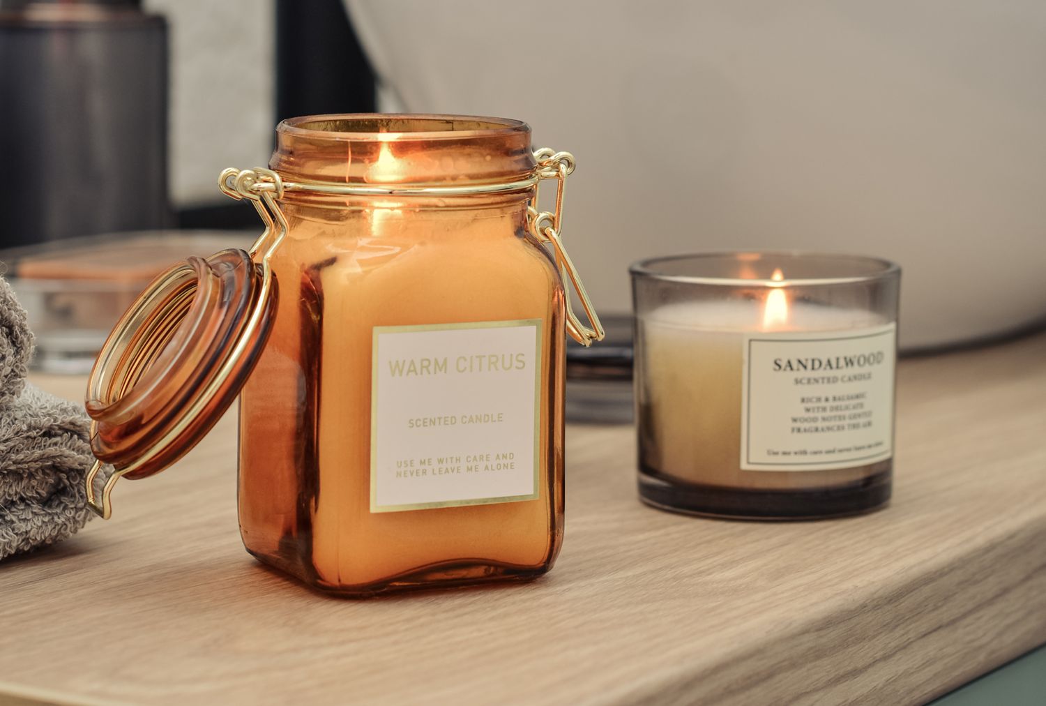 Two scented candles one in an orange jar and the other in a dark glass jar placed on a wooden bathroom counter