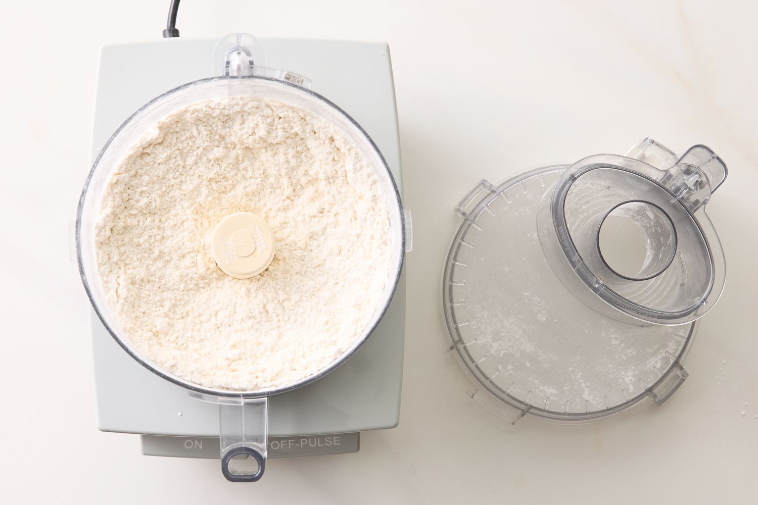 A food processor with cookie dough ingredients inside next to its lid on a countertop