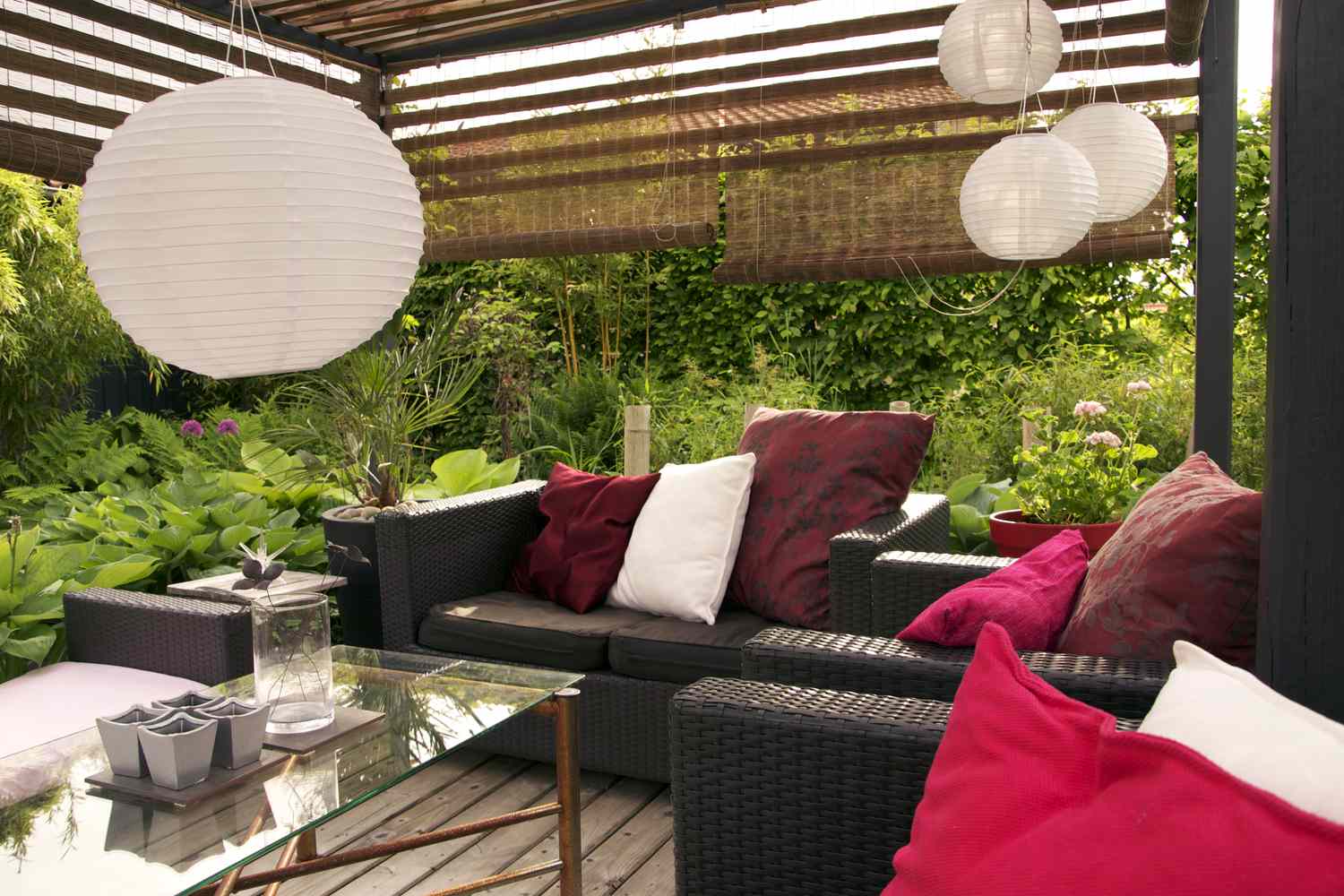 Outdoor patio with rattan furniture, cushions, and hanging lanterns, surrounded by lush greenery