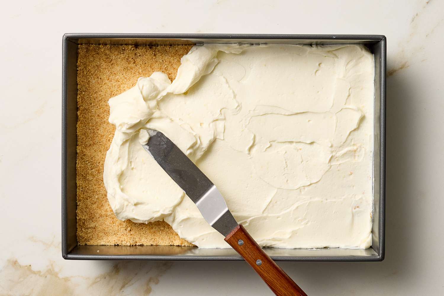 A rectangular pan with a graham cracker crust partially covered with cream mixture with a knife used for spreading the mixture