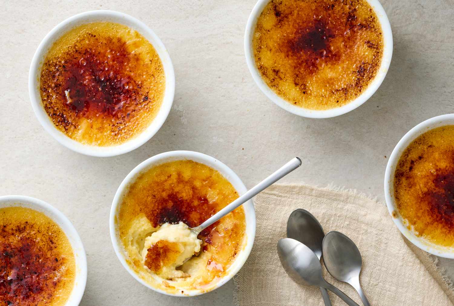 Multiple creme brulee servings in ramekins one partially eaten with spoons beside them on a cloth napkin