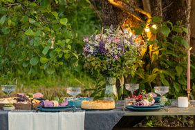 An outdoor table setting with flowers, plates, and glassware arranged in a natural setting under trees