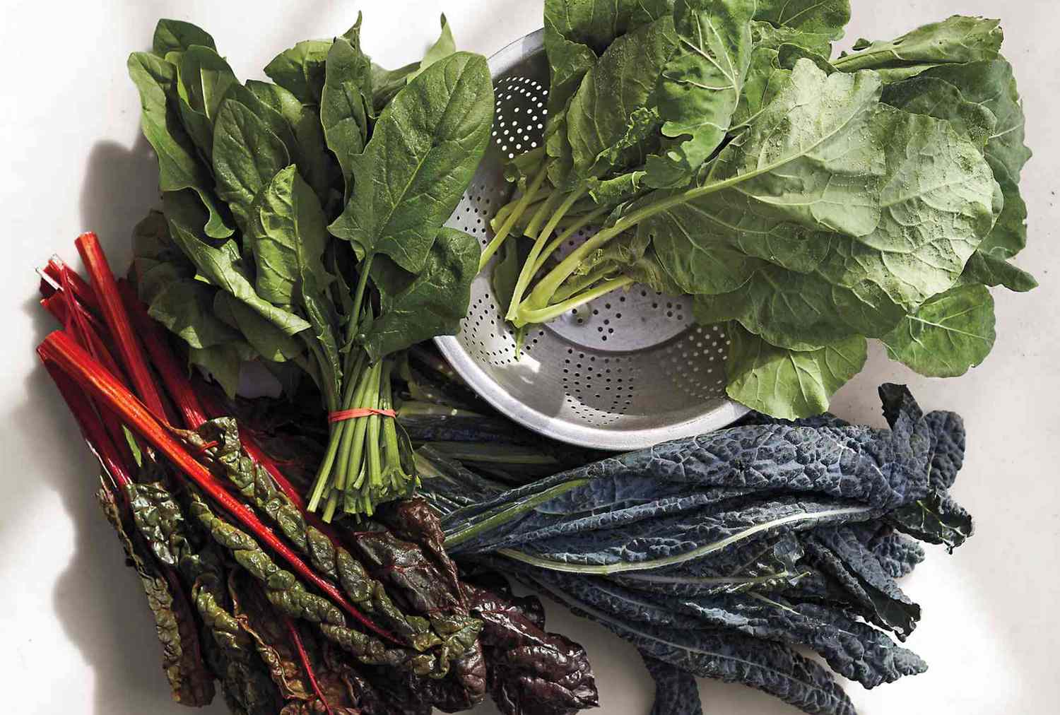 Various fresh leafy greens arranged on a surface including chard spinach kale and collard greens around a colander