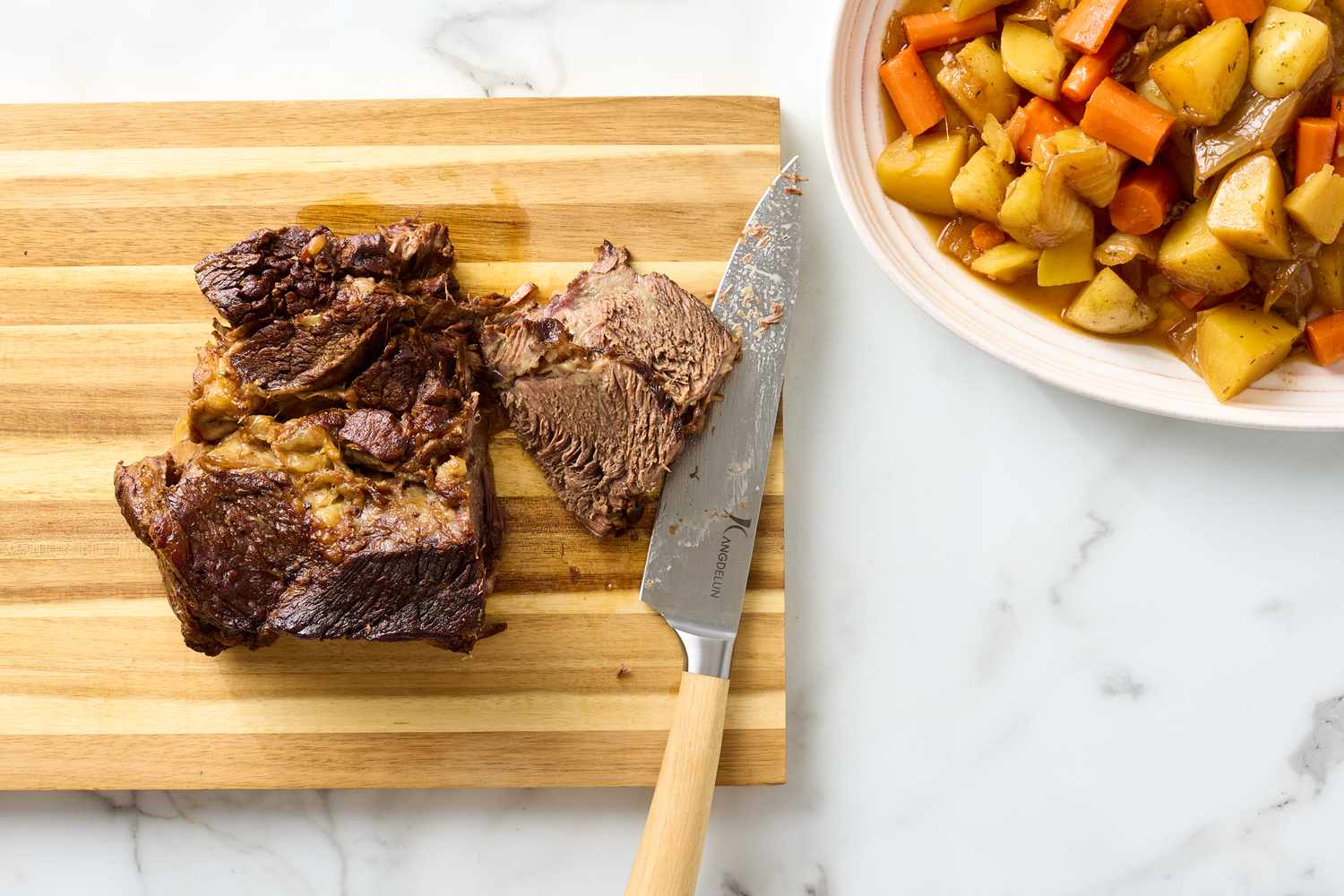 Cut pot roast on a wooden board with a knife and a bowl of vegetables nearby