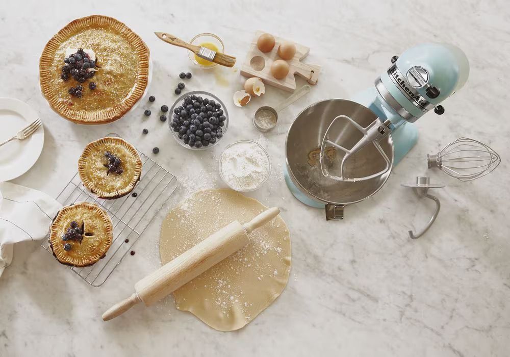 Kitchen setup with a stand mixer baked pies rolling pin and blueberries on a countertop