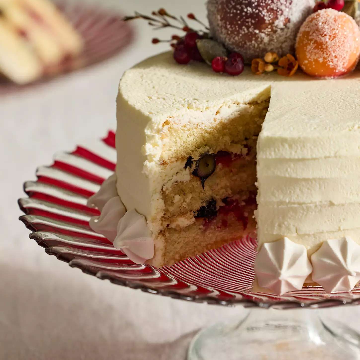 A sliced layer cake with frosting and decorated with berries and ornaments placed on a striped cake stand