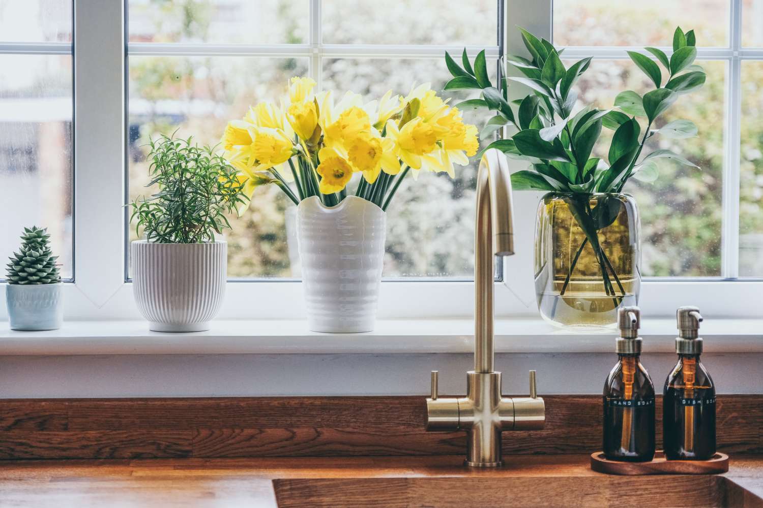 A kitchen window setup with potted plants daffodils in vases and soap dispensers on the countertop