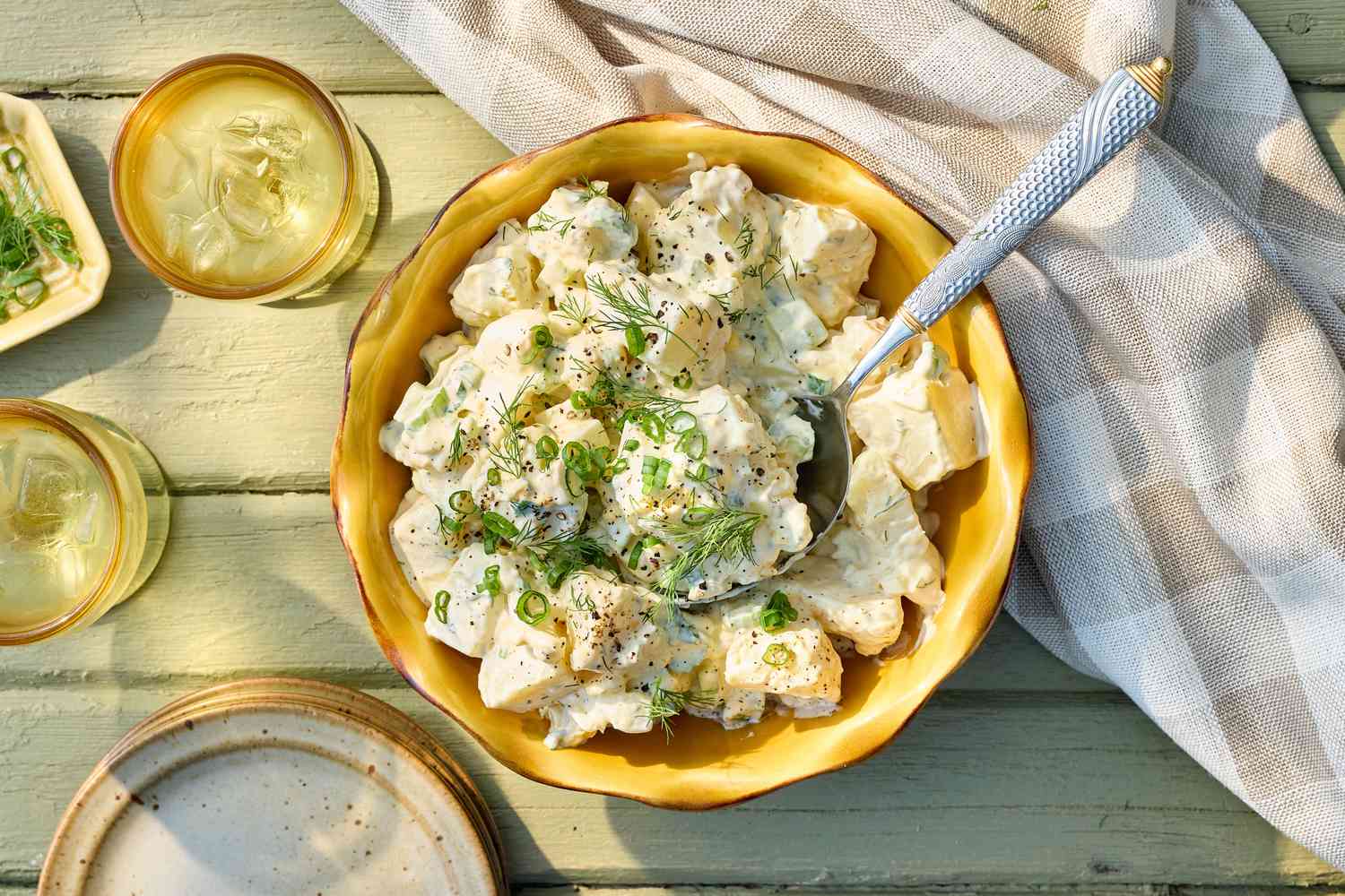 A bowl of classic potato salad garnished with green herbs, placed on a wooden table with a cloth napkin and drinks nearby