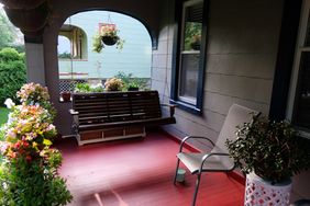 A porch with a swing bench a chair and potted plants viewed through an arched opening
