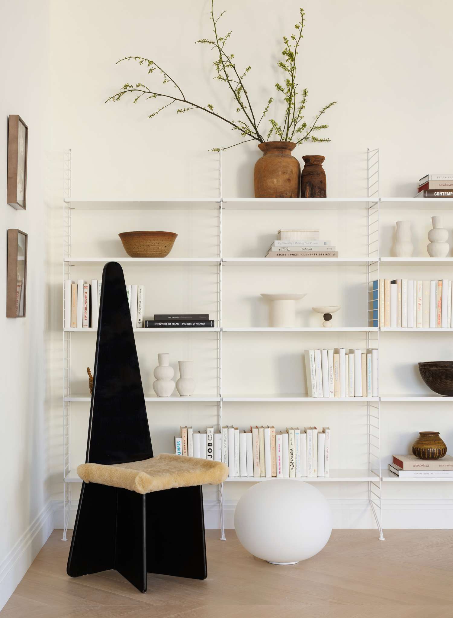 Minimalist living room by French California: black sculptural chair with sheepskin seat, white modular shelving with books, ceramics, and branches. White globe lamp, light wood floor, and large windows with natural light create a calm, stylish space.