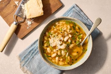 A bowl of bean soup topped with grated cheese and herbs placed on a napkin next to a cheese grater and block of cheese
