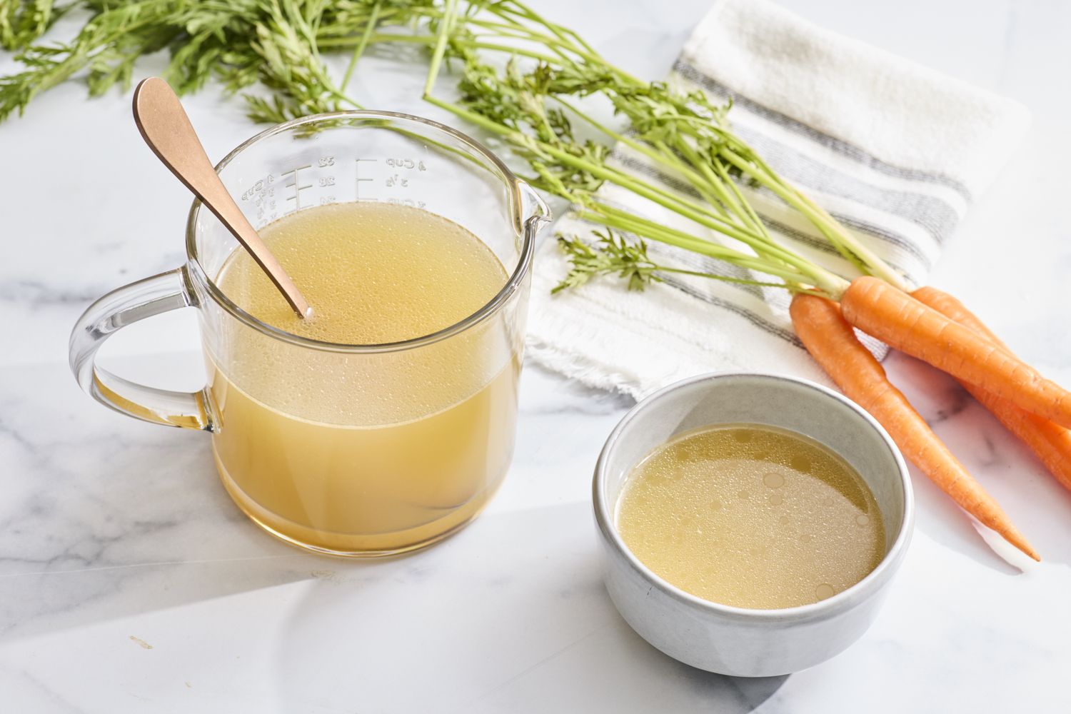A glass measuring cup and bowl containing chicken stock with fresh carrots and a cloth nearby