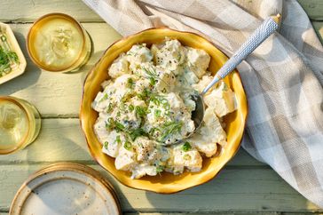 A bowl of classic potato salad garnished with green herbs, placed on a wooden table with a cloth napkin and drinks nearby