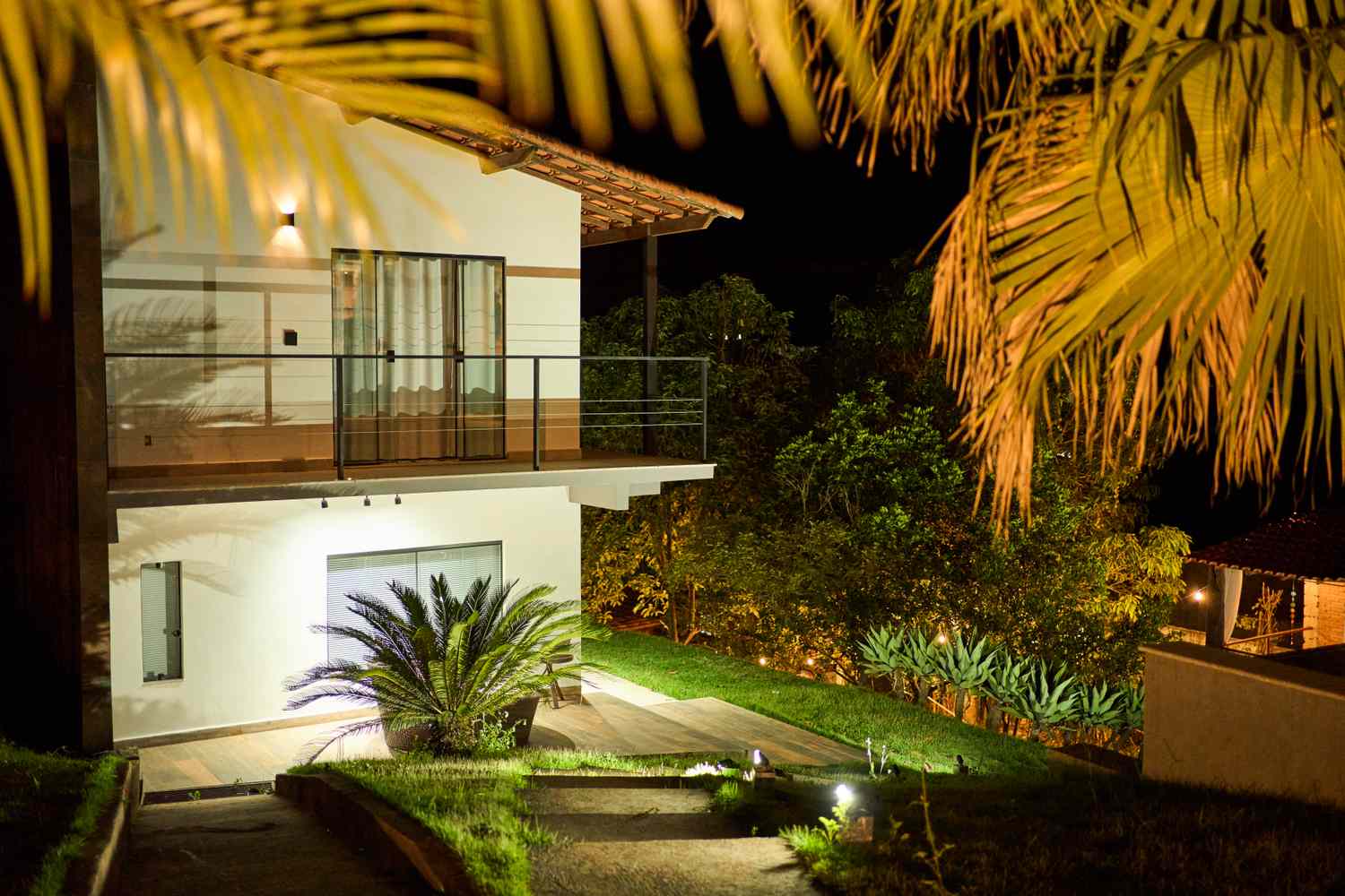 A modern twostory house at night surrounded by tropical plants and lit with outdoor lighting