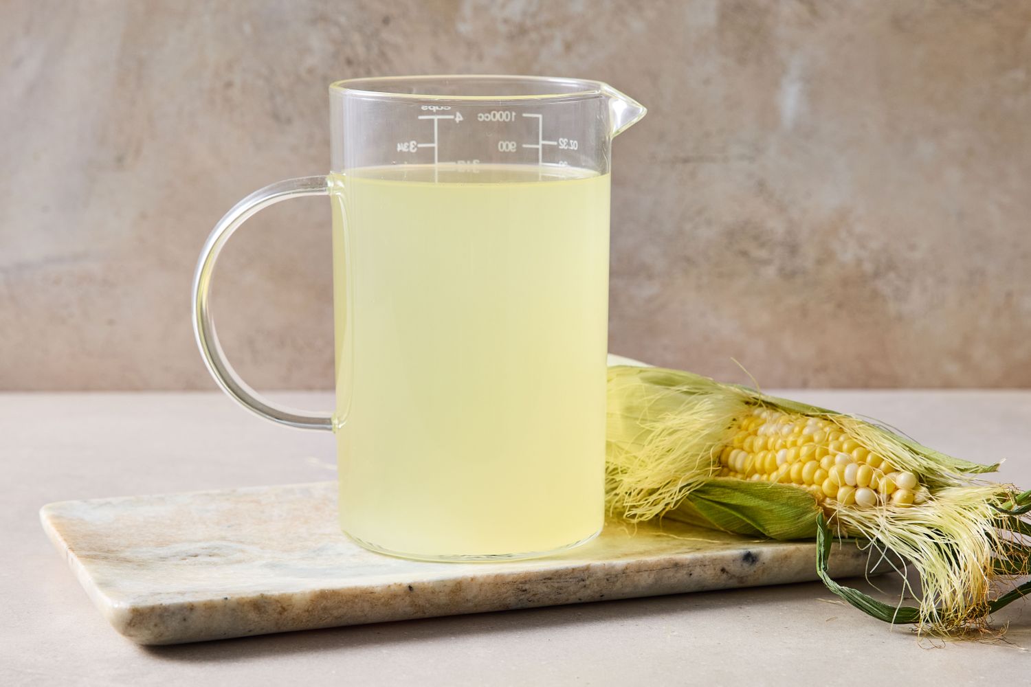 A glass measuring pitcher filled with liquid and a cob of corn on a cutting board