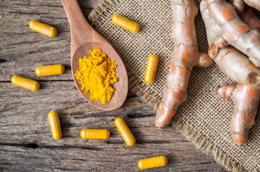Turmeric powder in a wooden spoon surrounded by capsules and turmeric roots on a rustic surface