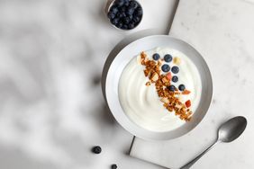 A bowl of yogurt topped with granola and blueberries a spoon and a small bowl of blueberries beside it