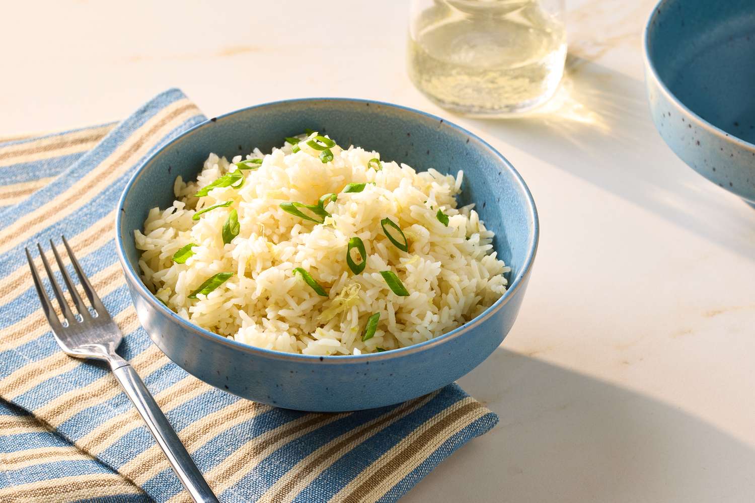 A bowl of rice garnished with green onions on a striped cloth alongside a fork