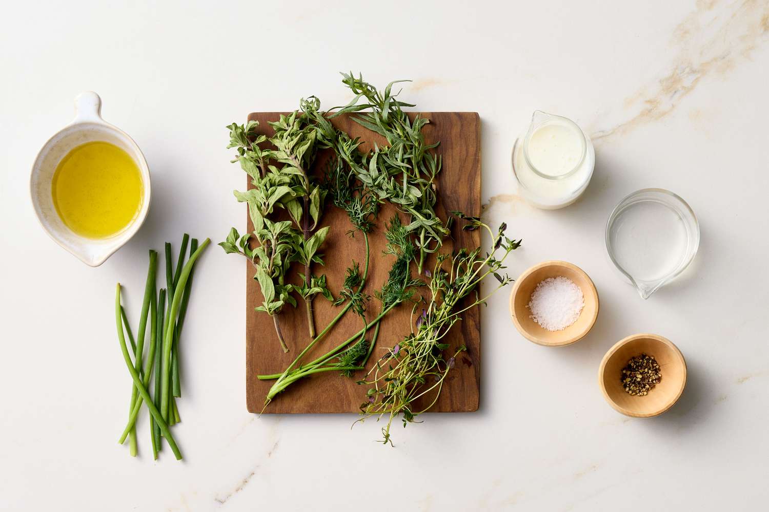 Ingredients for a buttermilk herb vinaigrette arranged on a white surface, including herbs, oil, buttermilk, salt, and pepper
