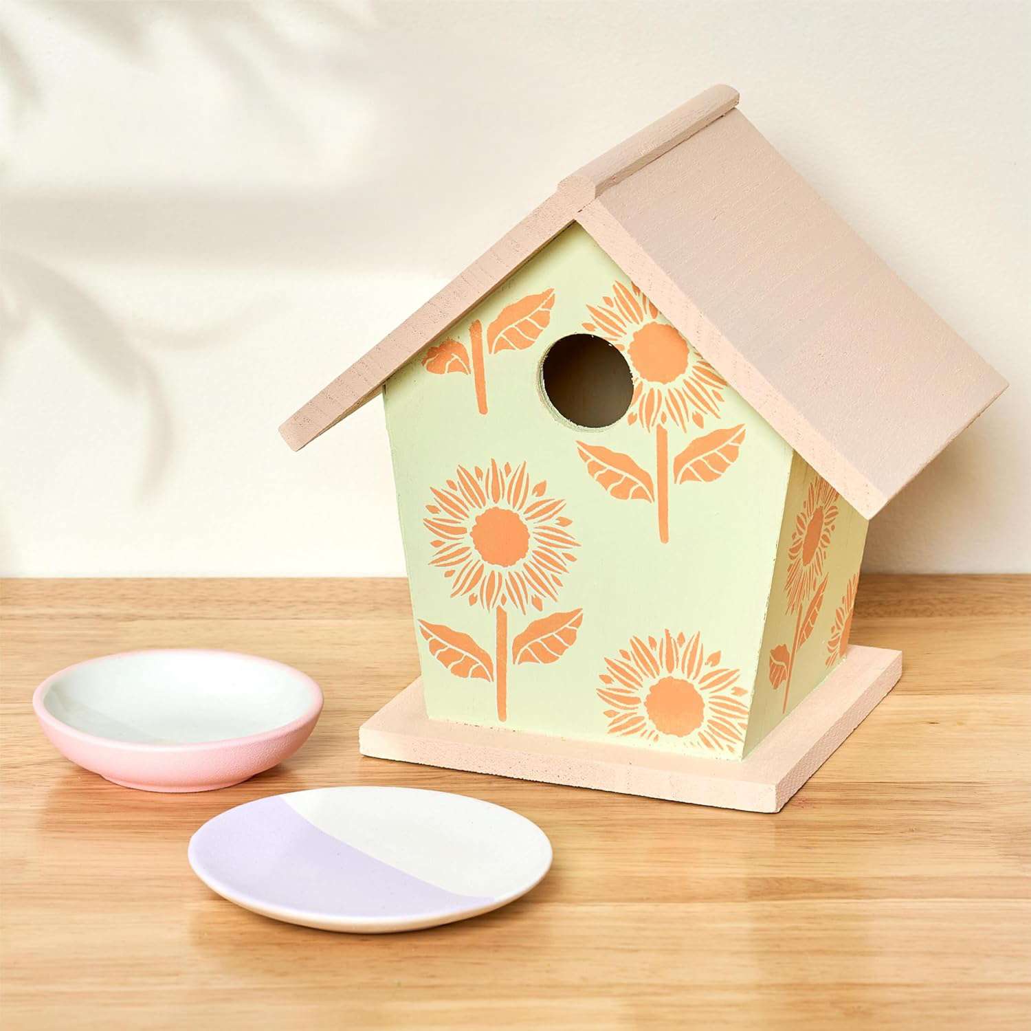 A birdhouse adorned with a sunflower design on a wooden table next to two small dishes