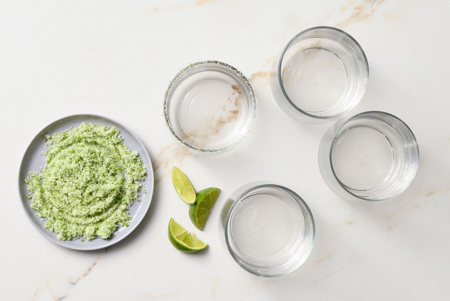 A flat lay setup of four glasses, a plate with green sugar, and lime wedges on a marble surface