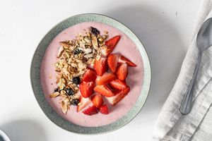 A smoothie bowl topped with granola and sliced strawberries placed on a table with a napkin and a spoon