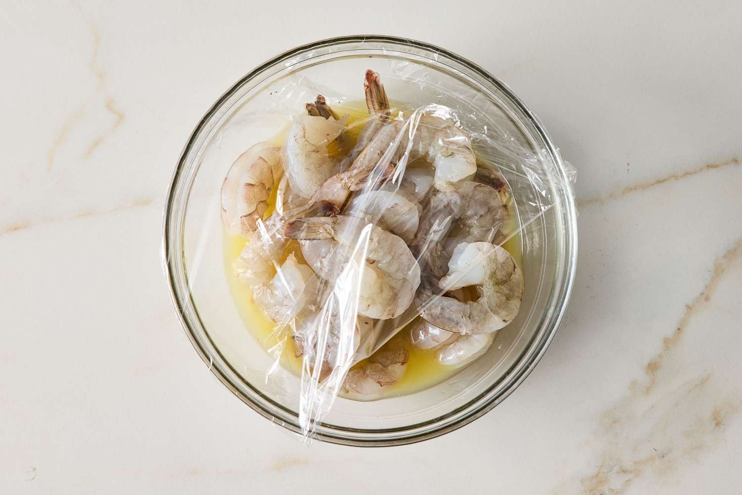 A bowl of raw shrimp covered with plastic wrap set on a lightcolored surface
