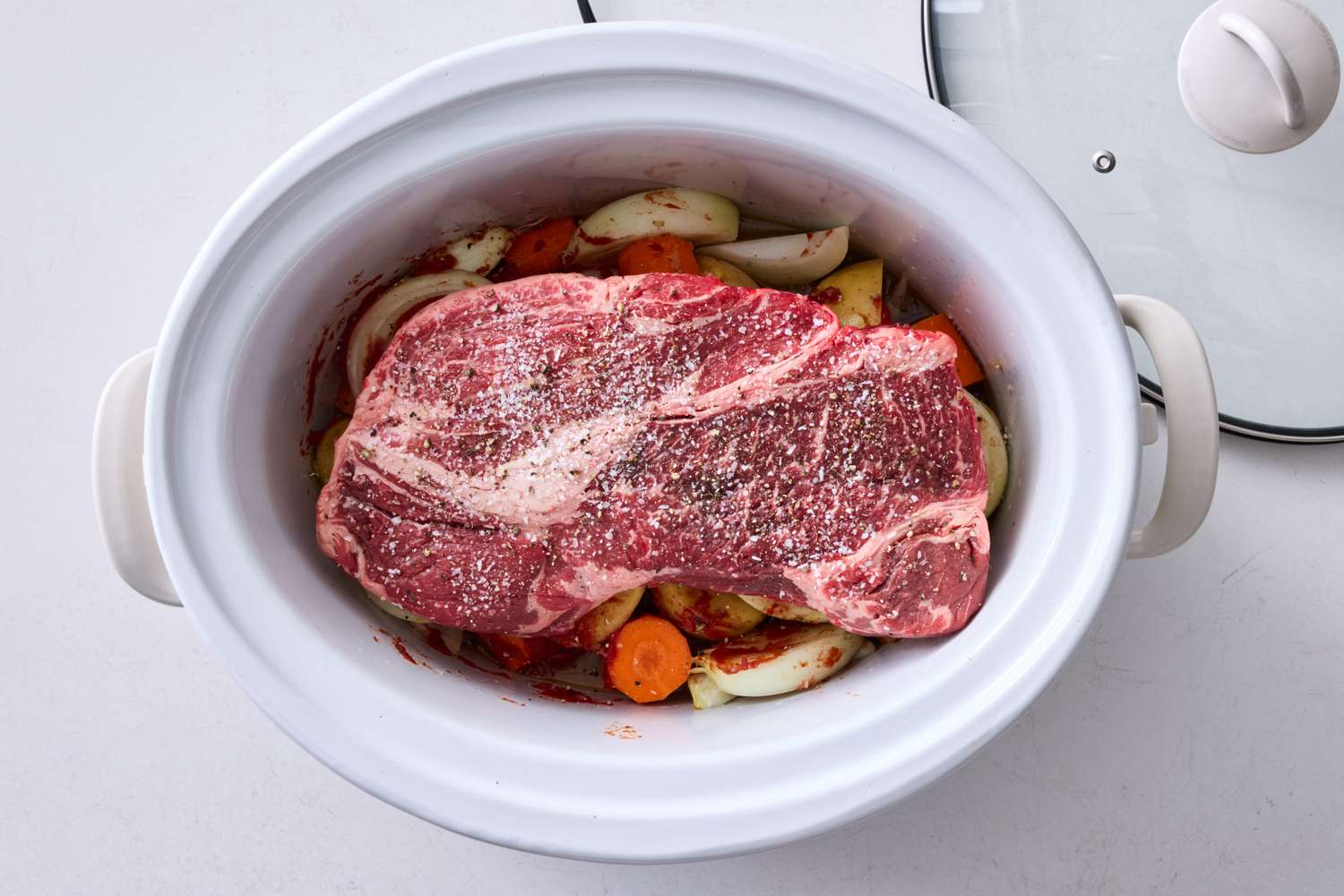 Uncooked seasoned pot roast with carrots and onions in a white crockpot