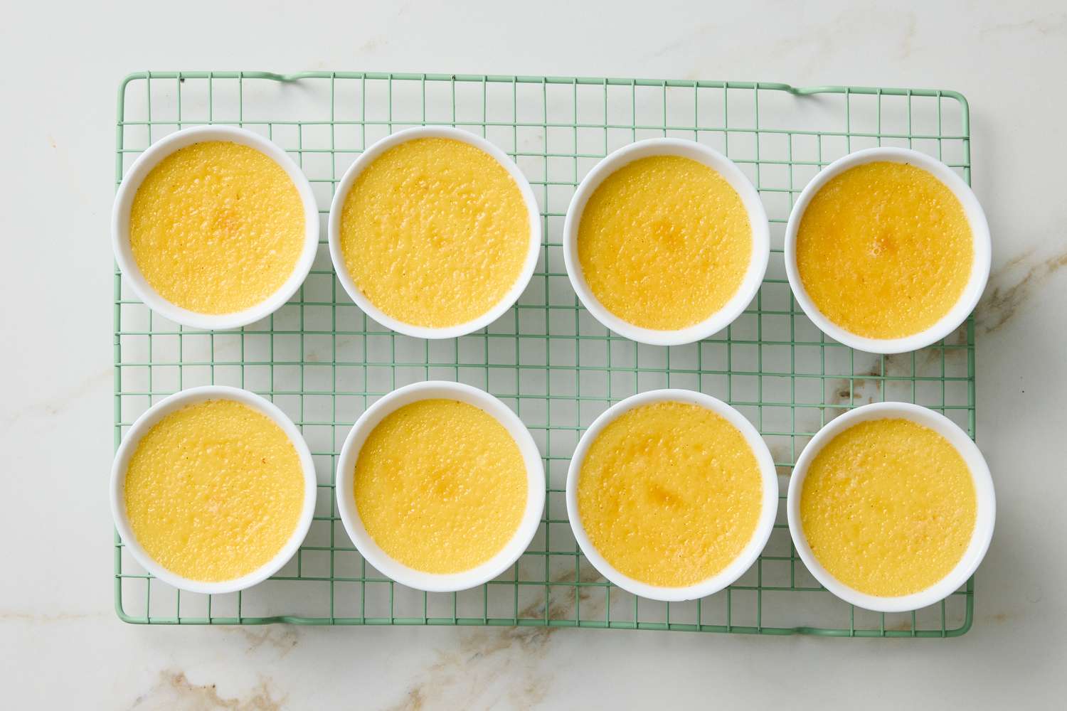 Eight creme brulee desserts in small white dishes arranged on a cooling rack