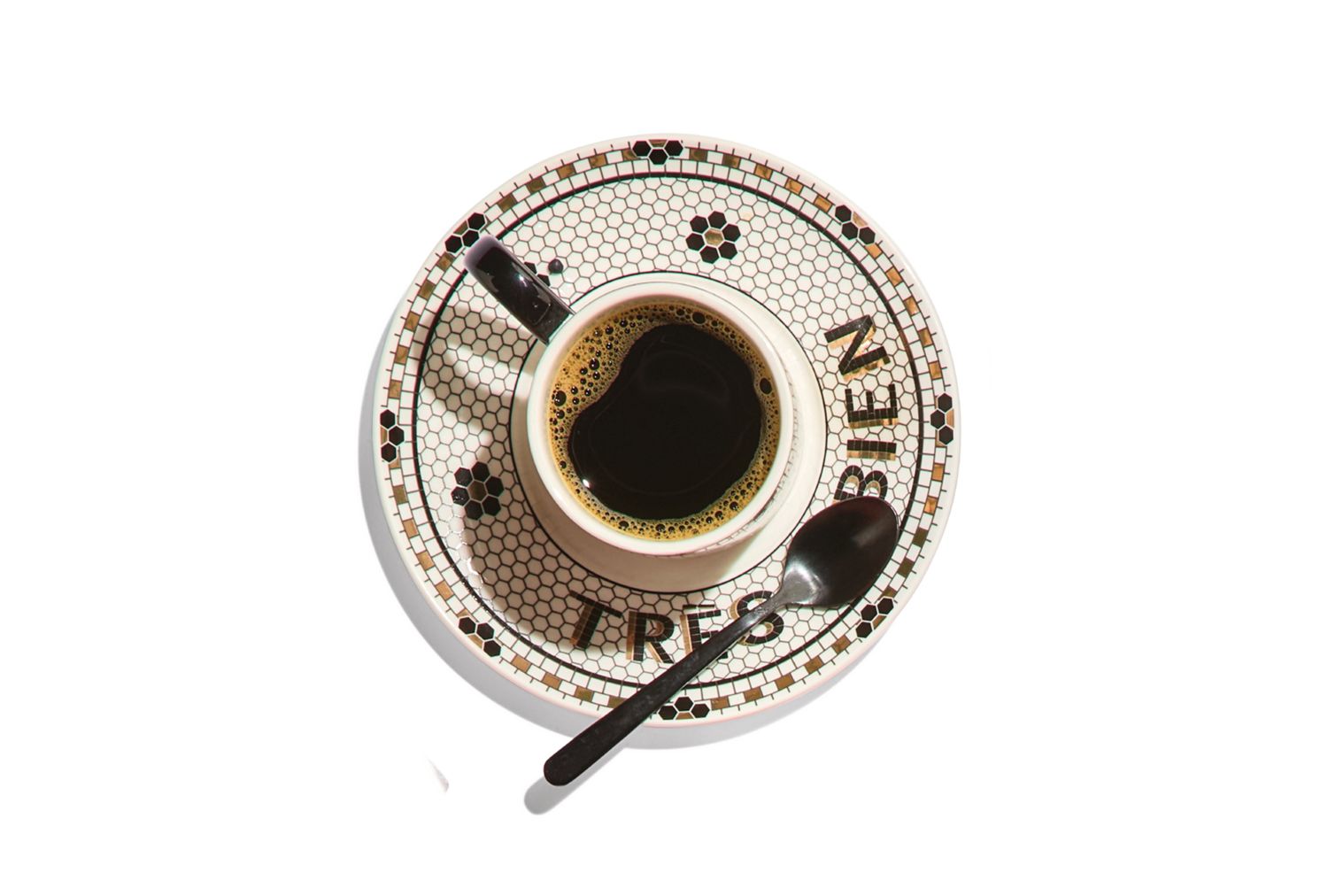 A cup of coffee on a patterned saucer with a spoon the saucer has Tres Bien written on it