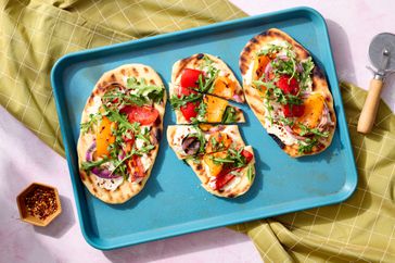 Grilled flatbread pizzas with toppings including roasted peppers, red onions, and arugula, tray and pizza cutter nearby