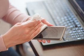 A person cleaning a smartphone with a cloth a laptop visible in the background
