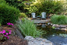 Two chairs by a backyard pond surrounded by plants and flowers