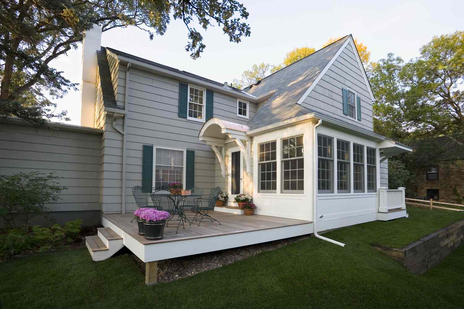 A house with a wooden deck and chairs with a potted plant surrounded by grass and trees in the background