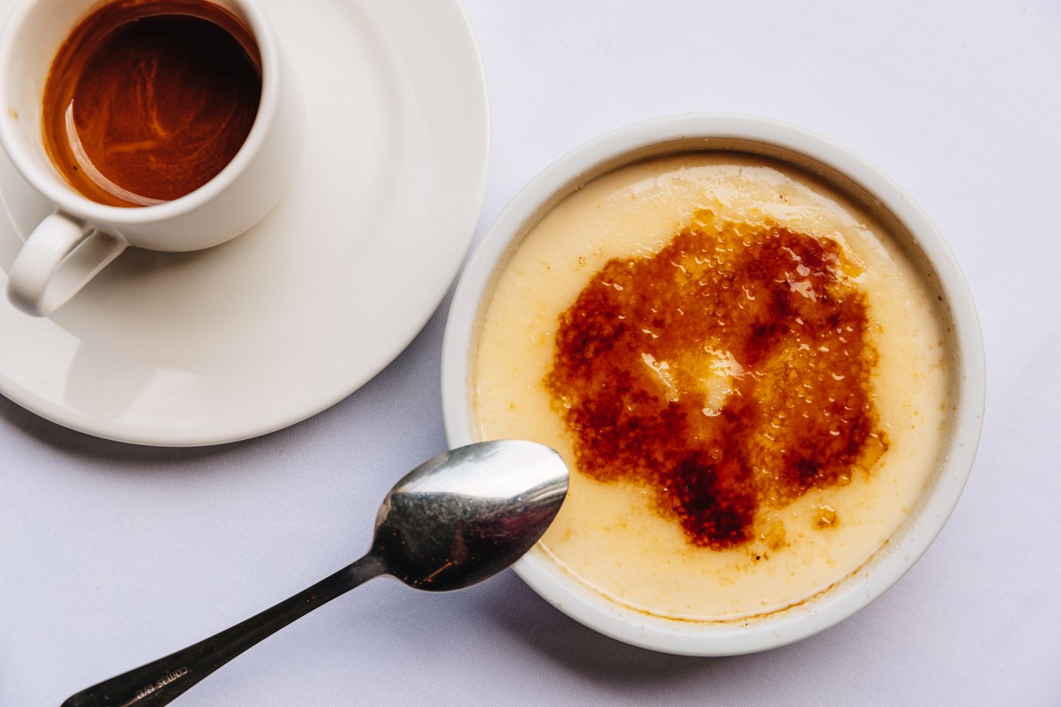 Crme brle with a spoon on a white surface beside a cup of espresso